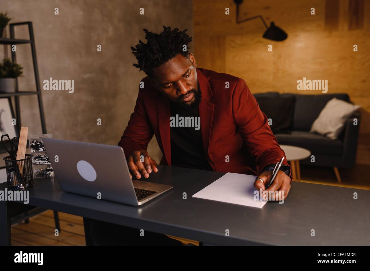 Bell'uomo nero afroamericano che lavora sul computer portatile mentre si siede dietro la scrivania in un accogliente soggiorno. Freelance che lavora da casa Foto Stock