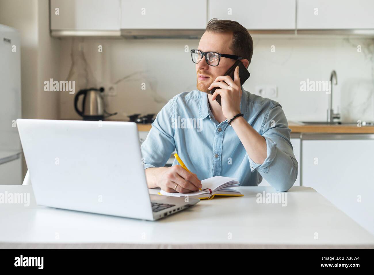 Sorridente freelance che lavora da casa, si siede alla scrivania della cucina di fronte al laptop, parlando sul telefono cellulare e prende appunti, multitasking uomo lavora a distanza Foto Stock