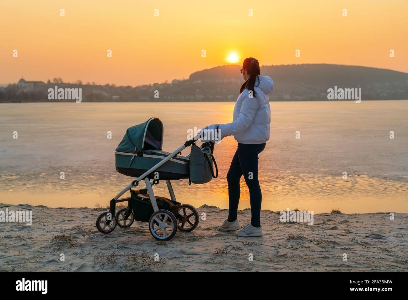 Una giovane madre cammina con un passeggino per un bambino sulla riva di un lago ghiacciato, al tramonto. Foto Stock
