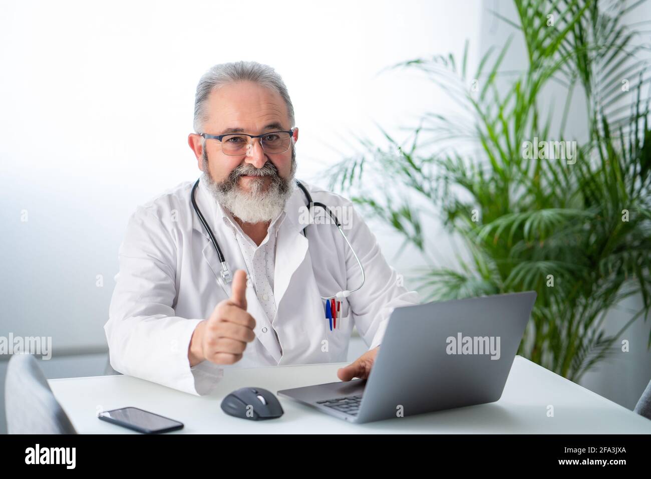 Medico anziano sopportato che lavora sorridendo con i pollici in su nel suo ufficio Foto Stock