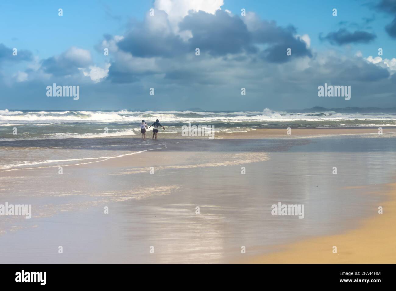 Due donne turisti - uno con le braccia aperte godono di levarsi in piedi fuori sulla spiaggia bagnata sotto il cielo tempestoso con l'oceano turbolento in sfondo Foto Stock