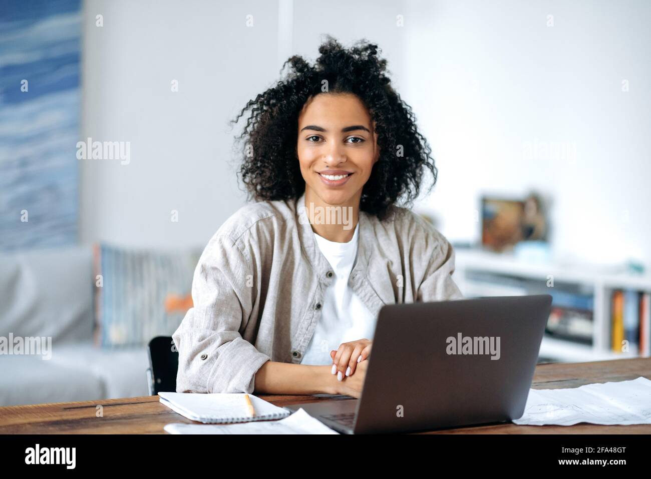 Bella amichevole giovane donna afro-americana sicuro in abiti casual di stile, freelancer, manager o studente seduto al computer portatile, di lavoro o di studio a casa, guardando la fotocamera, sorridendo Foto Stock