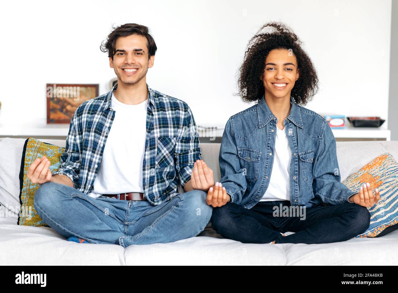 Coppia mista in amore passare il tempo insieme, rilassandosi e meditando mentre si siede in posizione lotus sul divano, alleviando lo stress, guardando la macchina fotografica, sorridendo. Ragazzo e la ragazza si prendono cura della loro salute Foto Stock