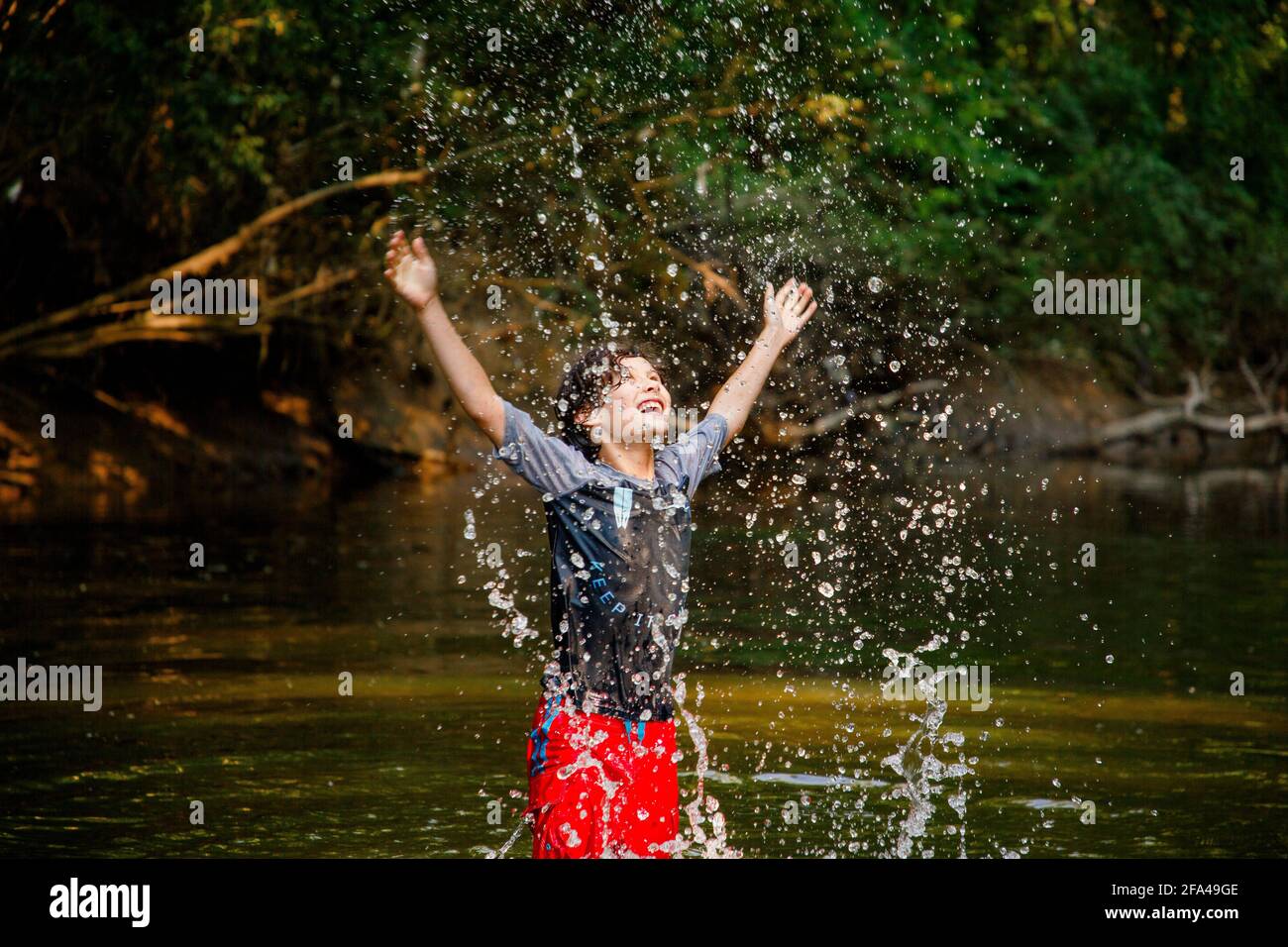 Un ragazzo gioioso che gioca nel fiume flings l'acqua cade in alto in aria Foto Stock