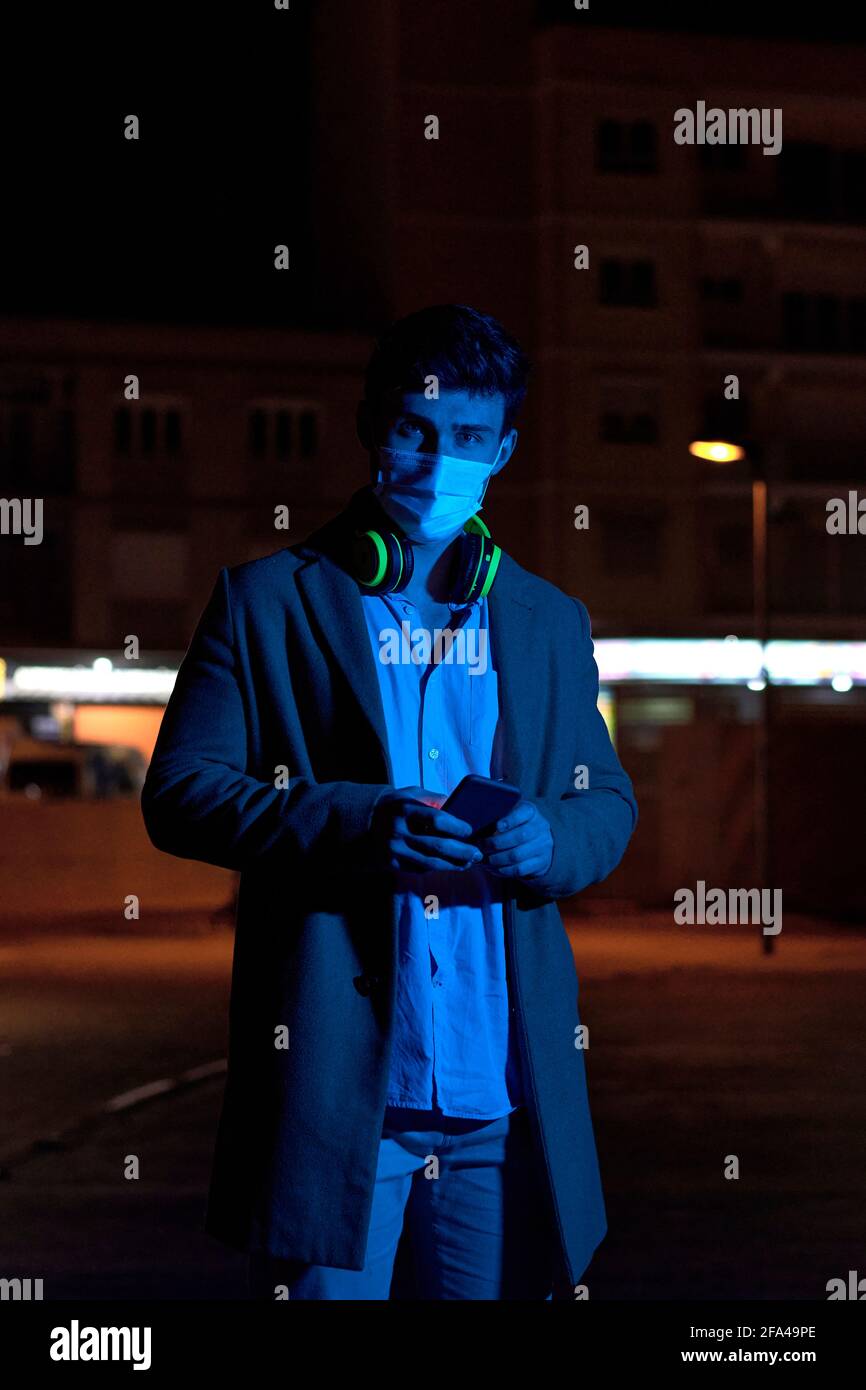 Giovane uomo d'affari con una maschera facciale e telefono all'aperto a. notte Foto Stock