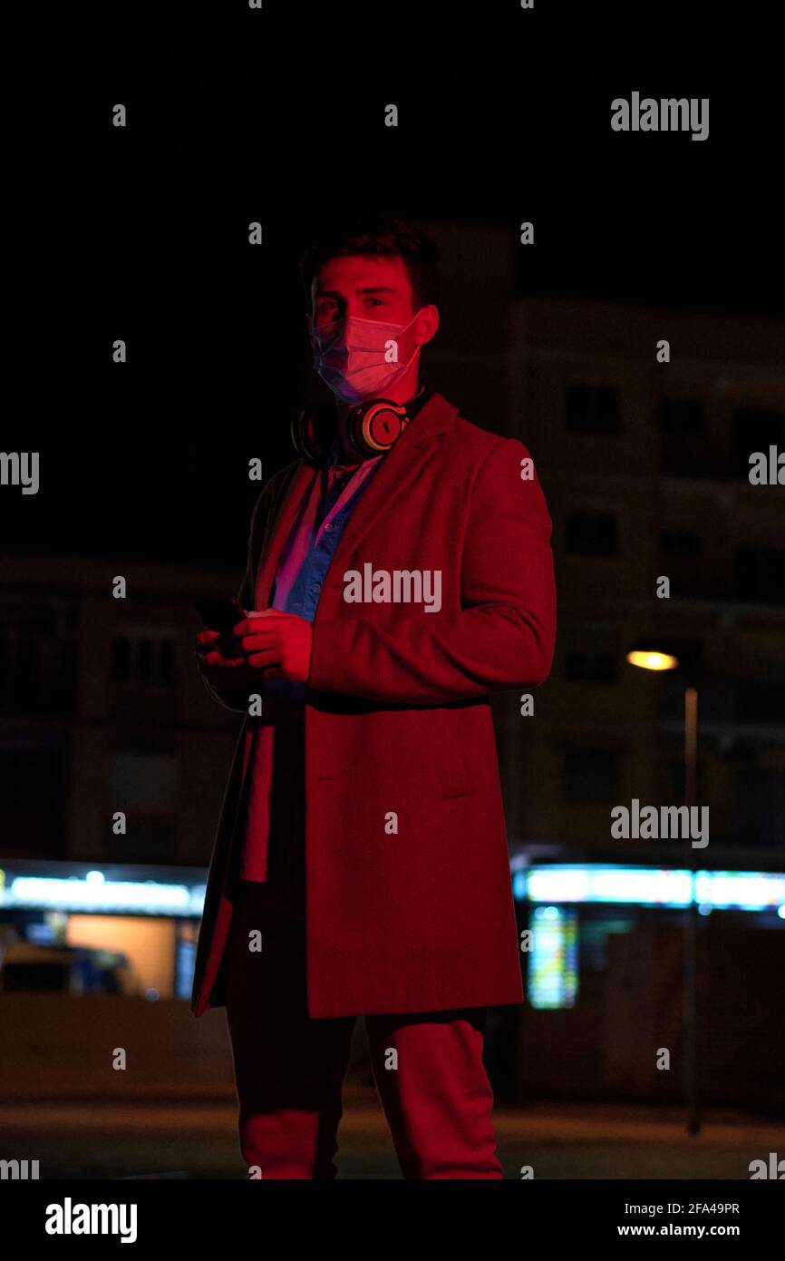 Giovane uomo d'affari con una maschera facciale e telefono all'aperto a. notte Foto Stock