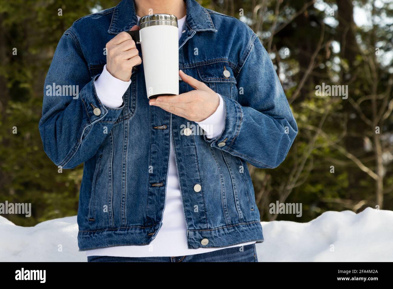 Mockup di una donna che indossa una giacca in denim che tiene una tazza da viaggio bianca da un bosco innevato. Tazza da caffè vuota da viaggio con dima per manico, modello mockup Foto Stock