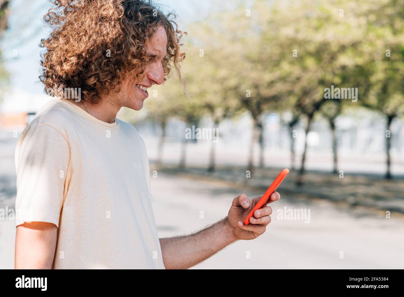 Ritratto orizzontale di un giovane dai capelli rossi che usa uno smartphone rosso sulla strada. Ha capelli ricci, vestiti casual e sorrisi Foto Stock