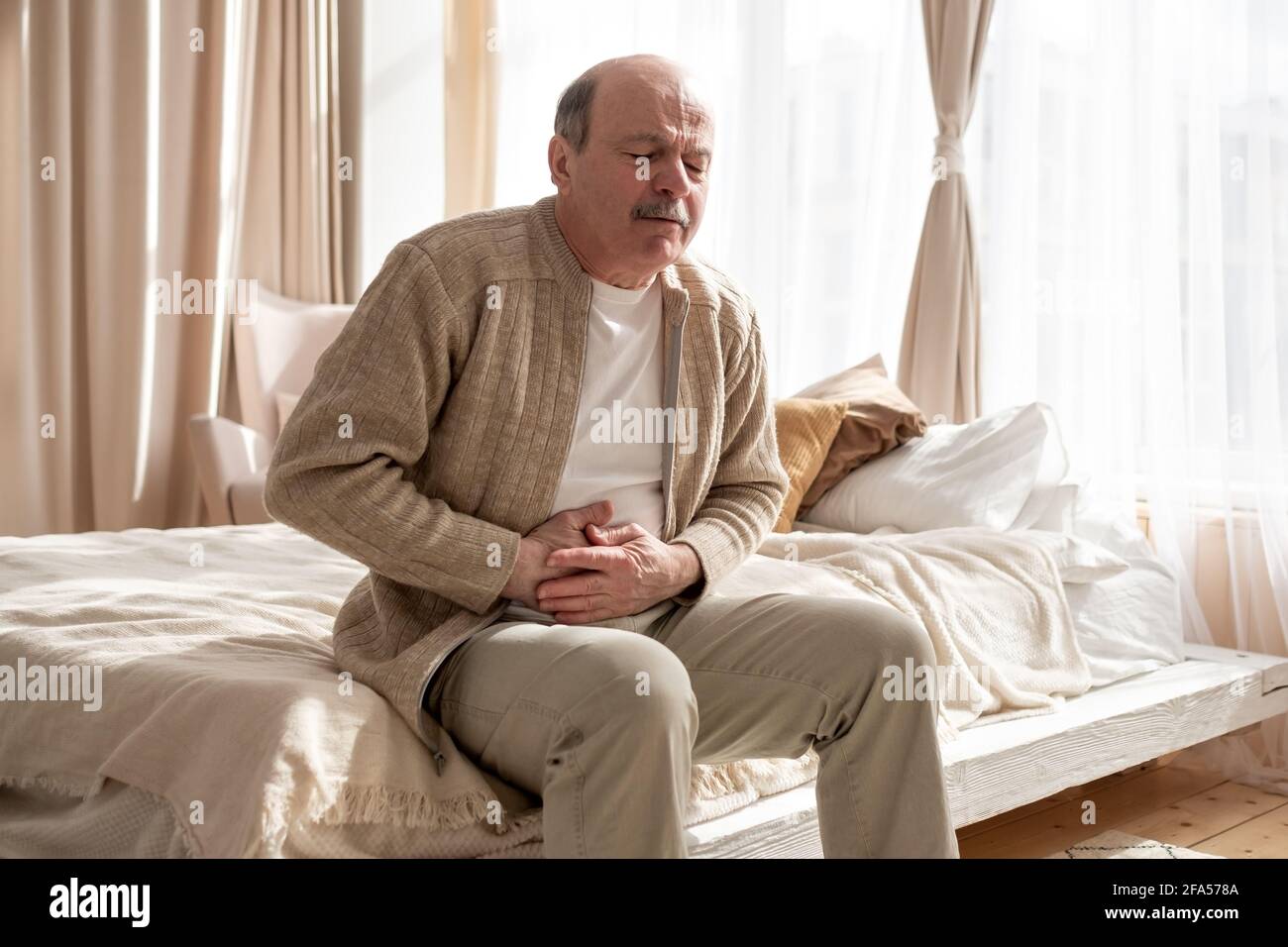 Uomo anziano che ha dolore dello stomaco seduto a casa. Nausea o dolore alla pancia. Foto Stock