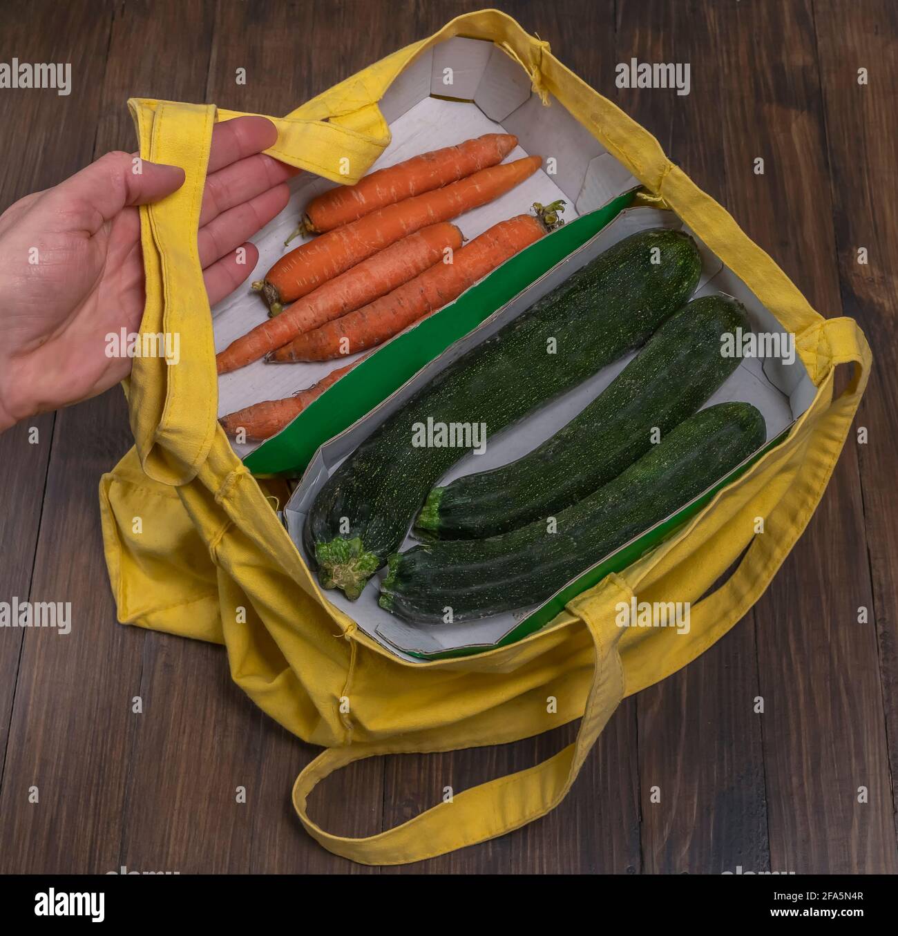 Una mano porta una borsa di tessuto giallo all'interno della quale sono presenti zucchine e carote su contenitori di cartone, su un tavolo di legno Foto Stock