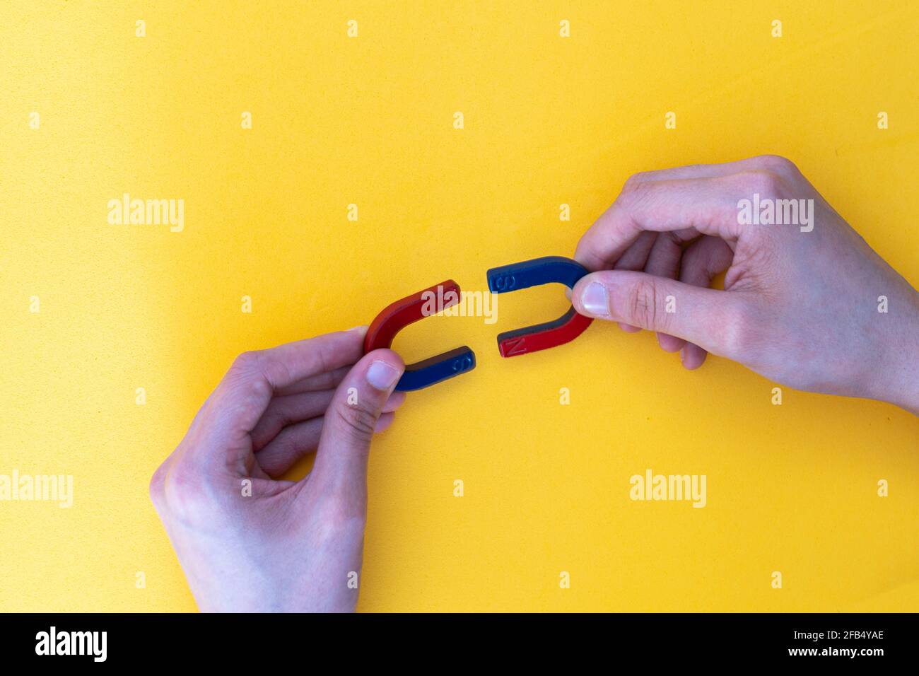 dettaglio delle mani che tengono due magneti blu e rosso a forma di ferro di cavallo che mostrano come i poli positivo e negativo sono uniti su uno sfondo giallo, conce Foto Stock