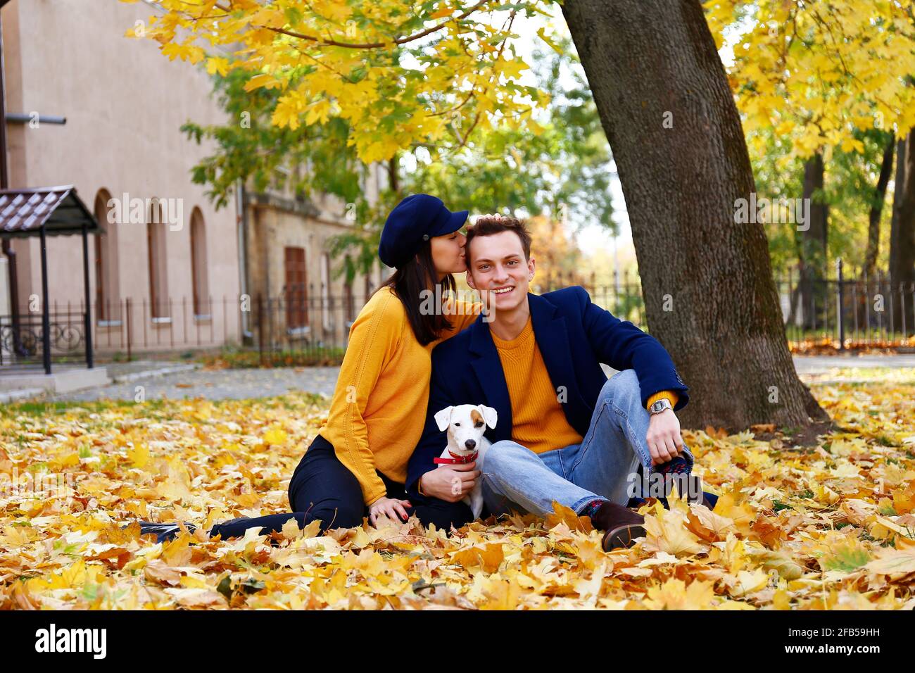 Attraente giovane coppia seduta su mucchio di foglie caduti nel mese di novembre con il loro cucciolo Jack Russell Terrier, foglie gialle a terra. Sfondo, c. Foto Stock