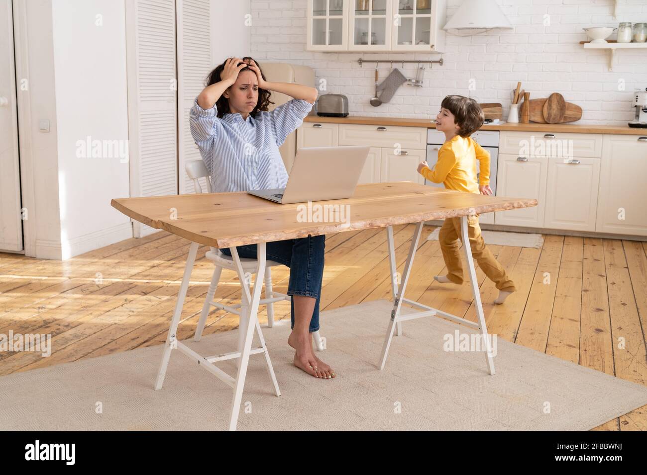 Donna lavoratrice freelance stanca di lavoro figlio rumoroso su computer portatile computer in cucina con un piccolo figlio in esecuzione Foto Stock