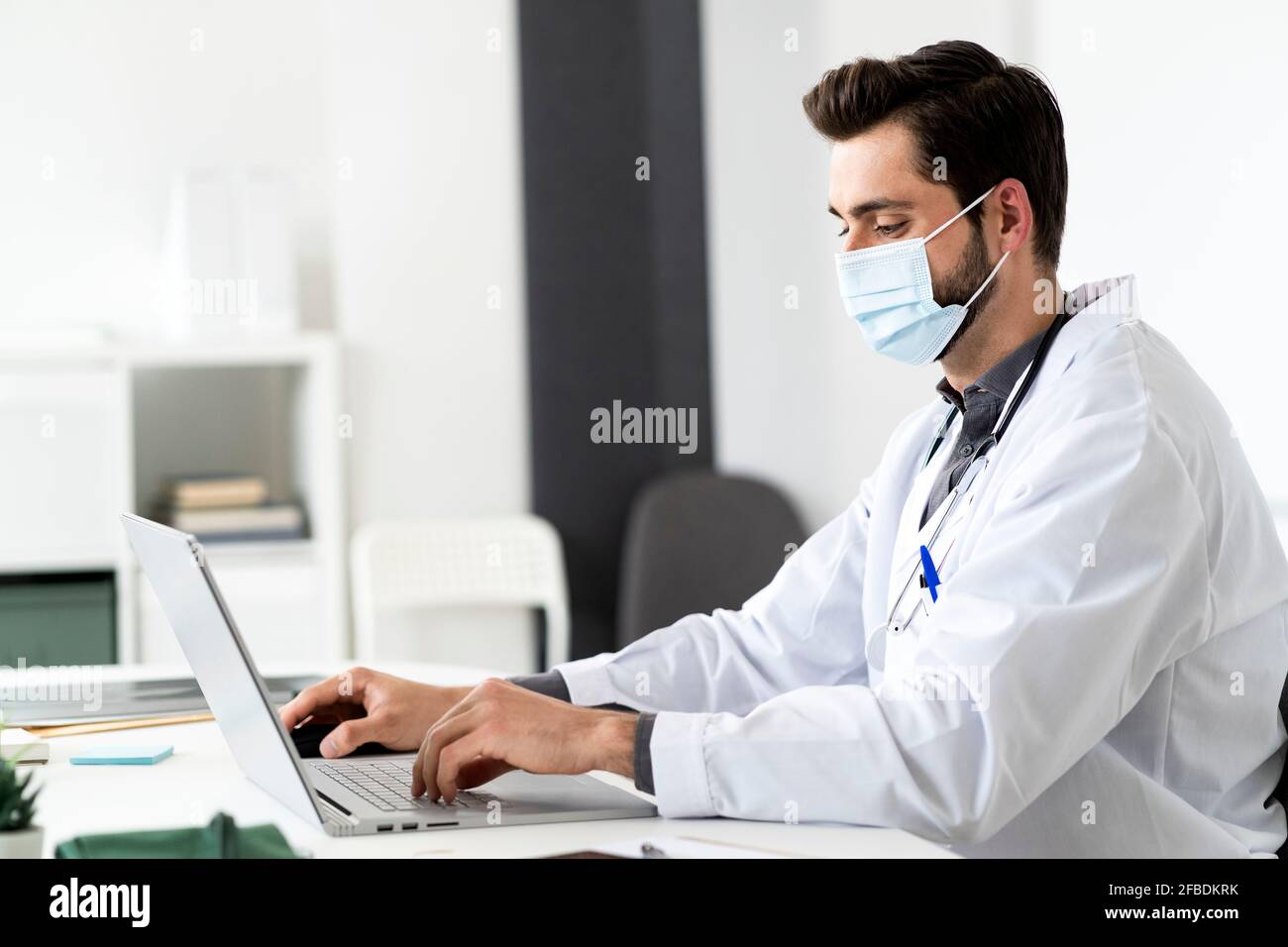 Medico di sesso maschile indossando la maschera mentre si usa il computer portatile alla scrivania Foto Stock