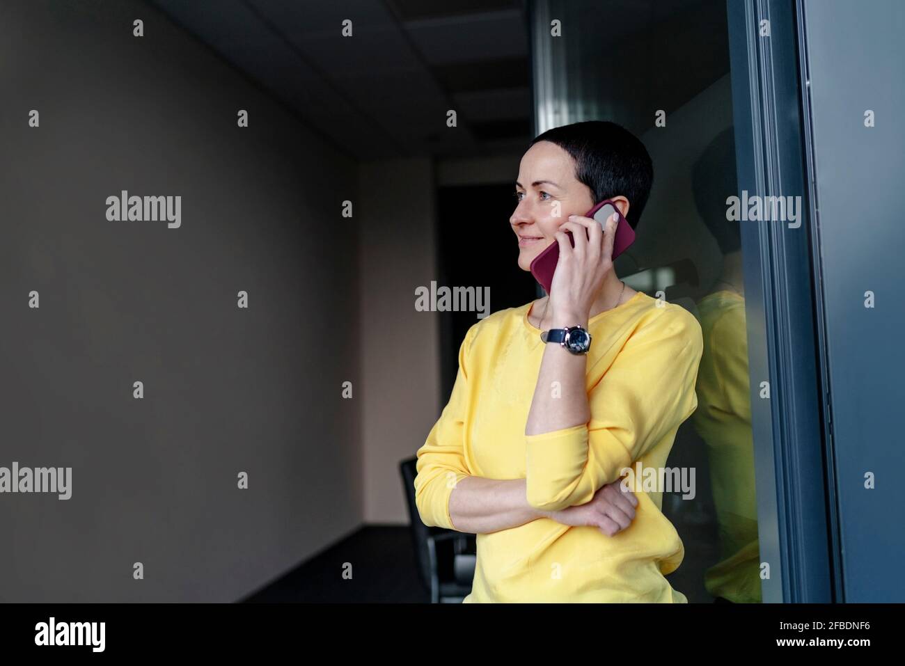 Donna d'affari sorridente che contempla mentre parla su smartphone in moderno ufficio Foto Stock