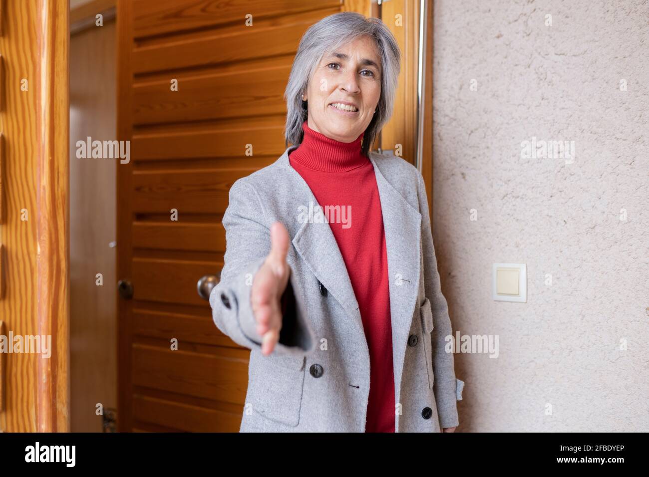 Donna sorridente in giacca che offre la stretta di mano sulla porta anteriore di casa Foto Stock