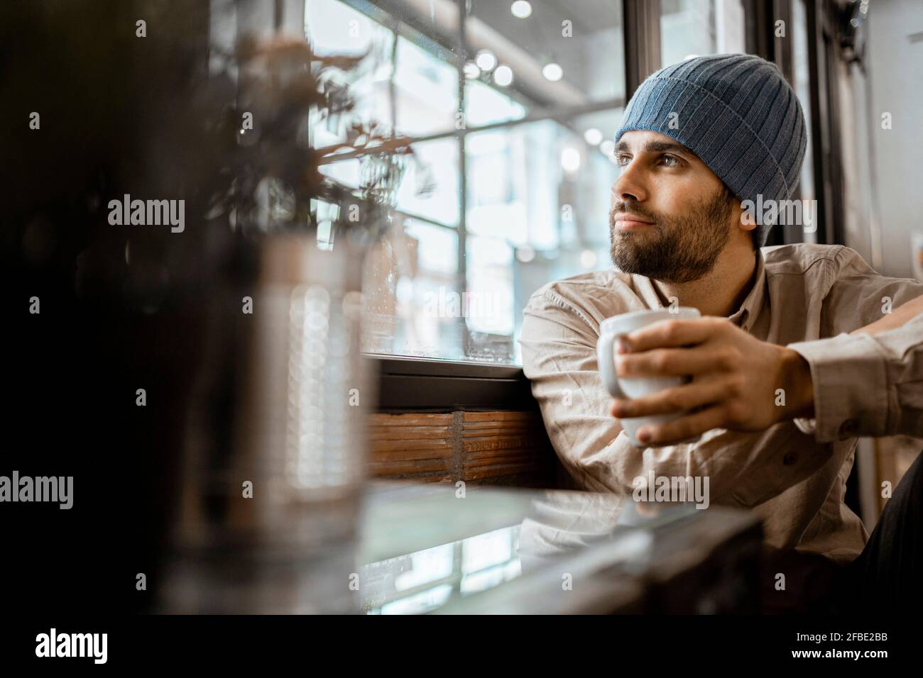 Uomo mid adult che indossa un cappello a maglia con una tazza di caffè attraverso la finestra nel caffè Foto Stock