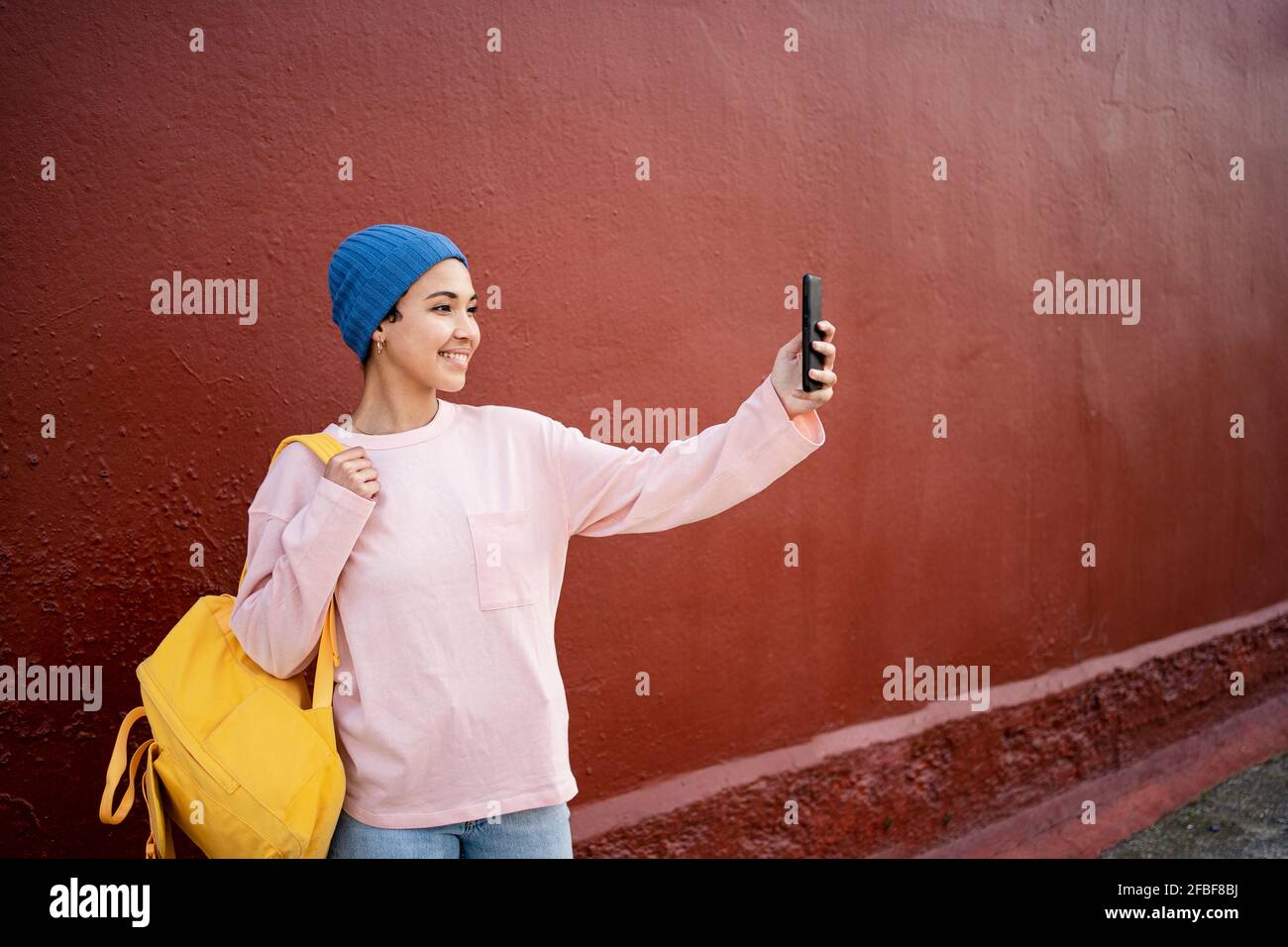 Donna sorridente che trasporta lo zaino mentre prende il selfie attraverso il telefono mobile davanti alla parete rossa Foto Stock