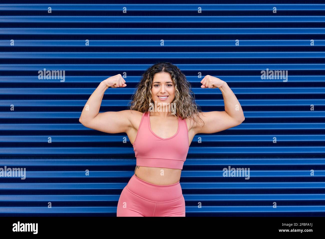 Femmina atleta sorridente che flette i muscoli davanti al corrugato blu parete Foto Stock