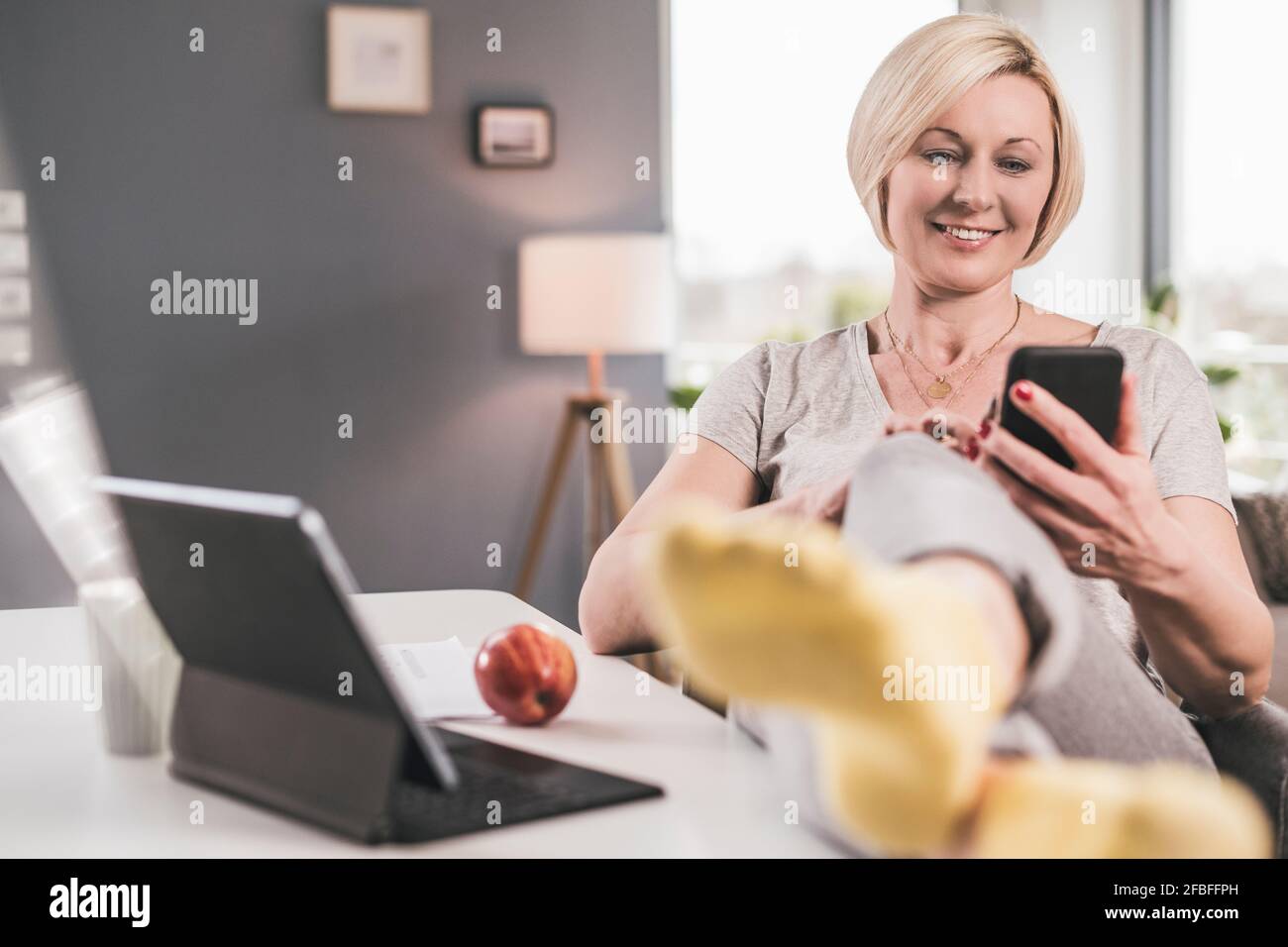 Donna sorridente che usa lo smartphone mentre si appoggiano le gambe sul tavolo a casa Foto Stock