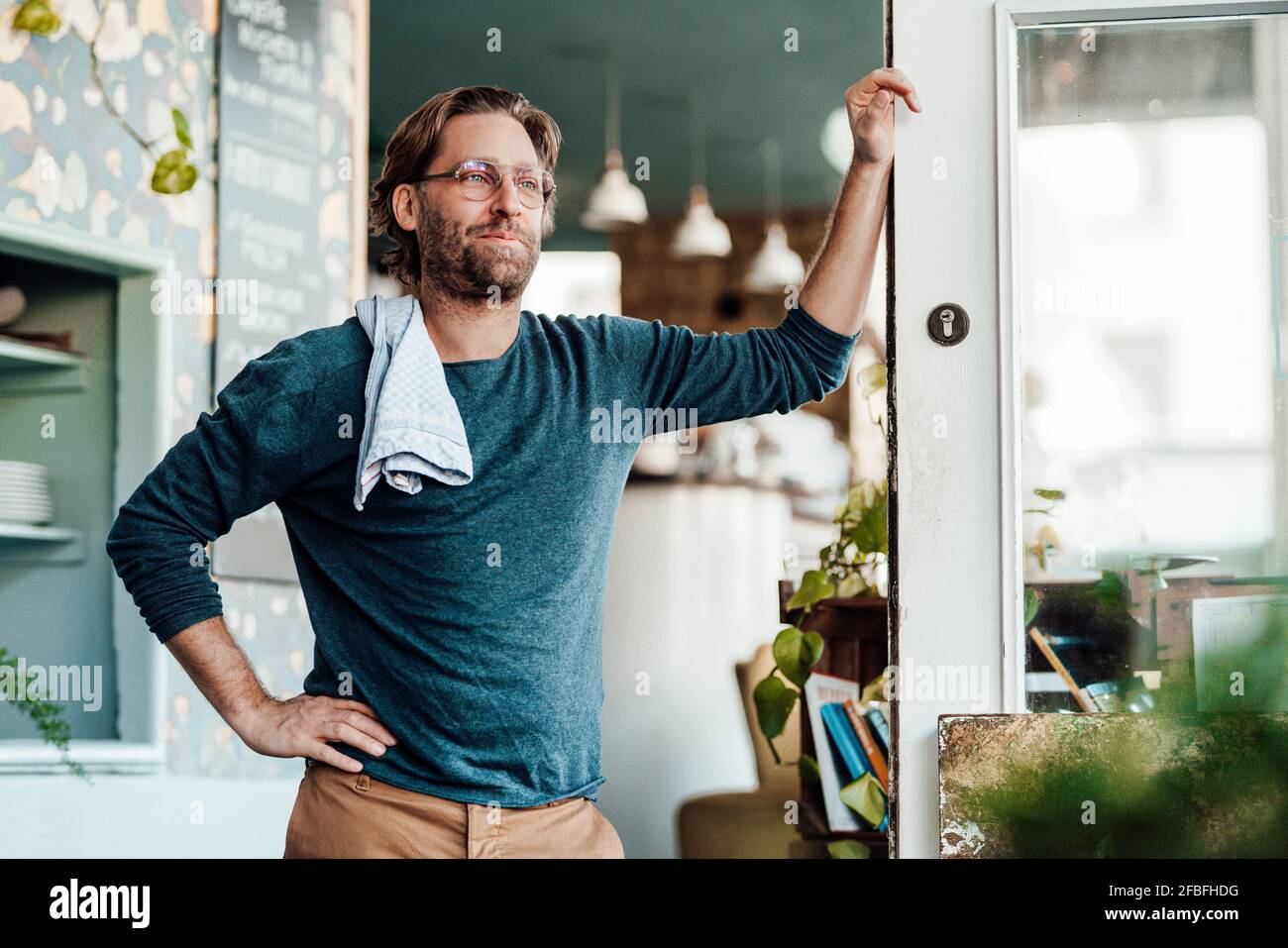 Proprietario maschile maturo con mano sull'anca appoggiata sulla porta al caffè Foto Stock