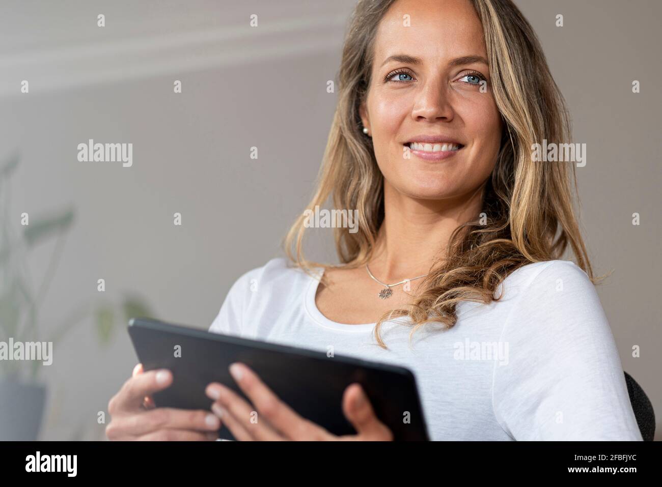 Donna sorridente con gli occhi blu che guardano via mentre tiene il digitale tablet a casa Foto Stock