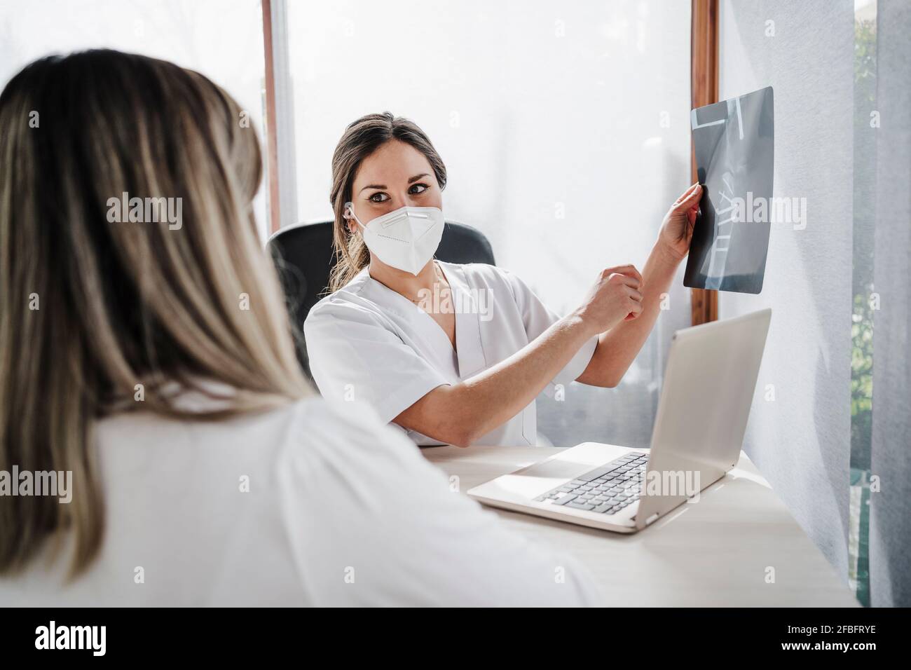 Medico femminile che guarda il paziente mentre tiene i raggi X medici vicino Laptop in clinica durante COVID-19 Foto Stock