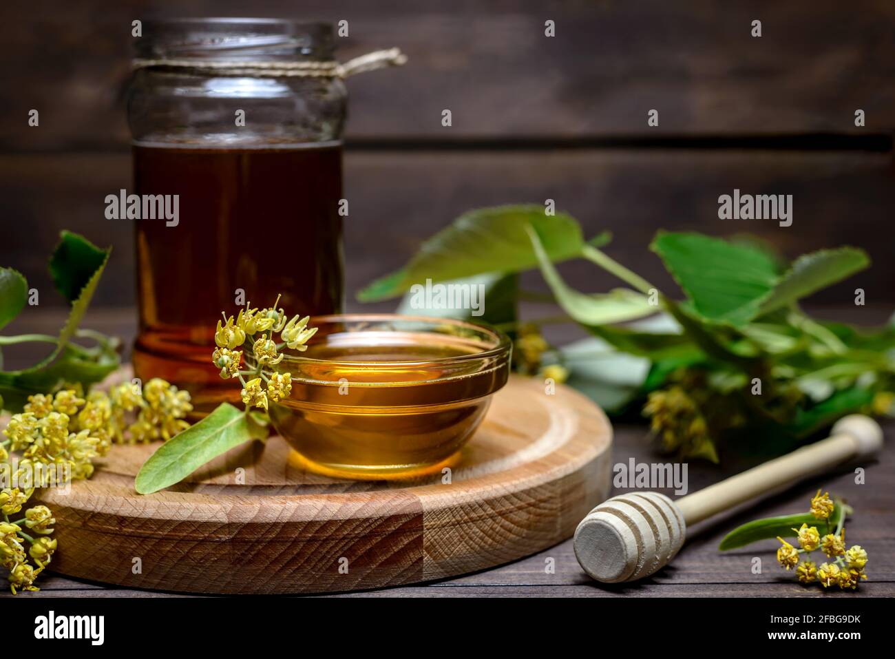 Vasetto e ciotola con miele di tiglio con bastone per miele e fiori di tiglio freschi su un tavolo di legno. Foto Stock