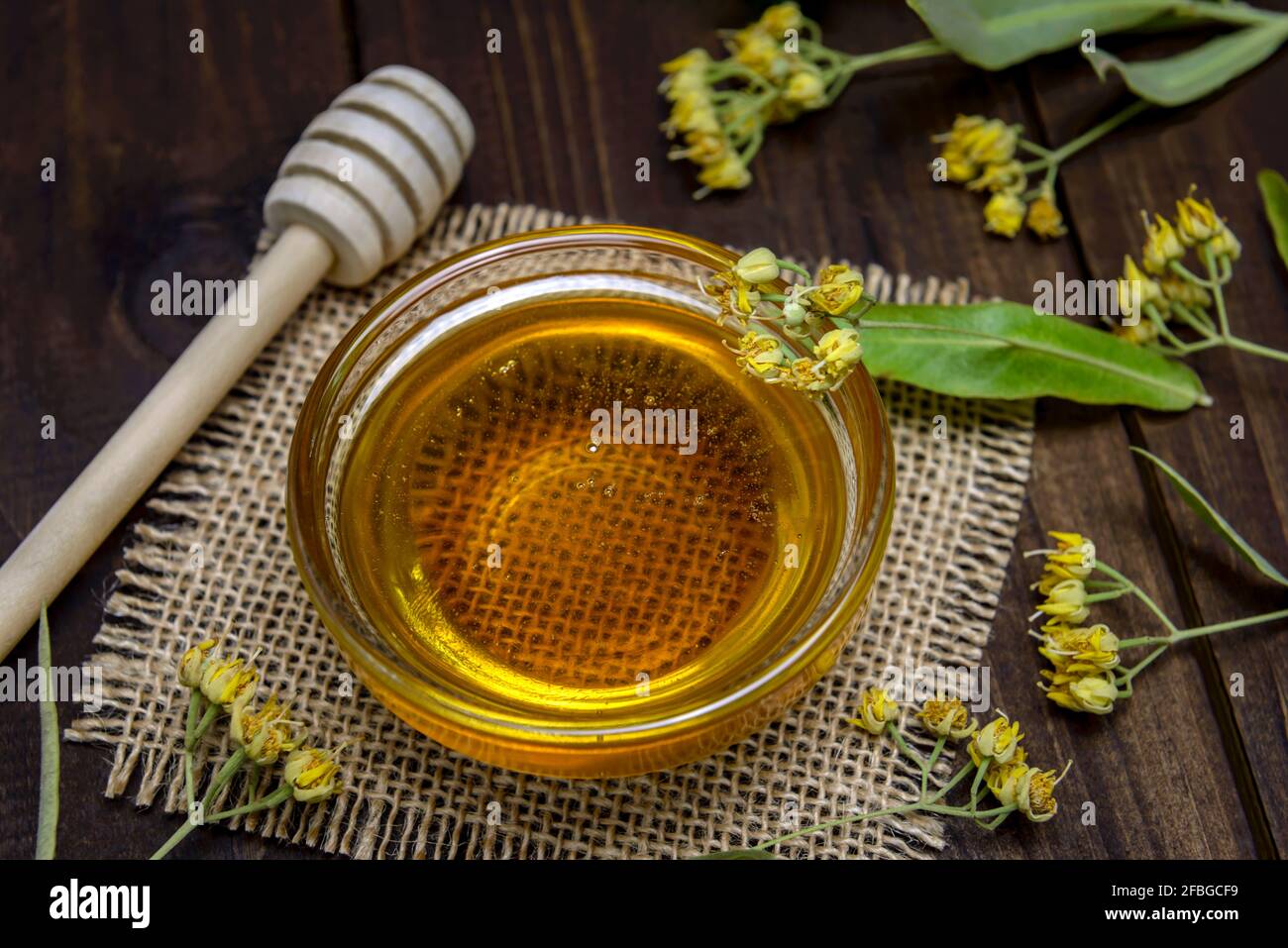 Ciotola di vetro con miele di tiglio, bastone per miele e fiori di tiglio freschi su un tavolo di legno. Messa a fuoco selettiva. Foto Stock