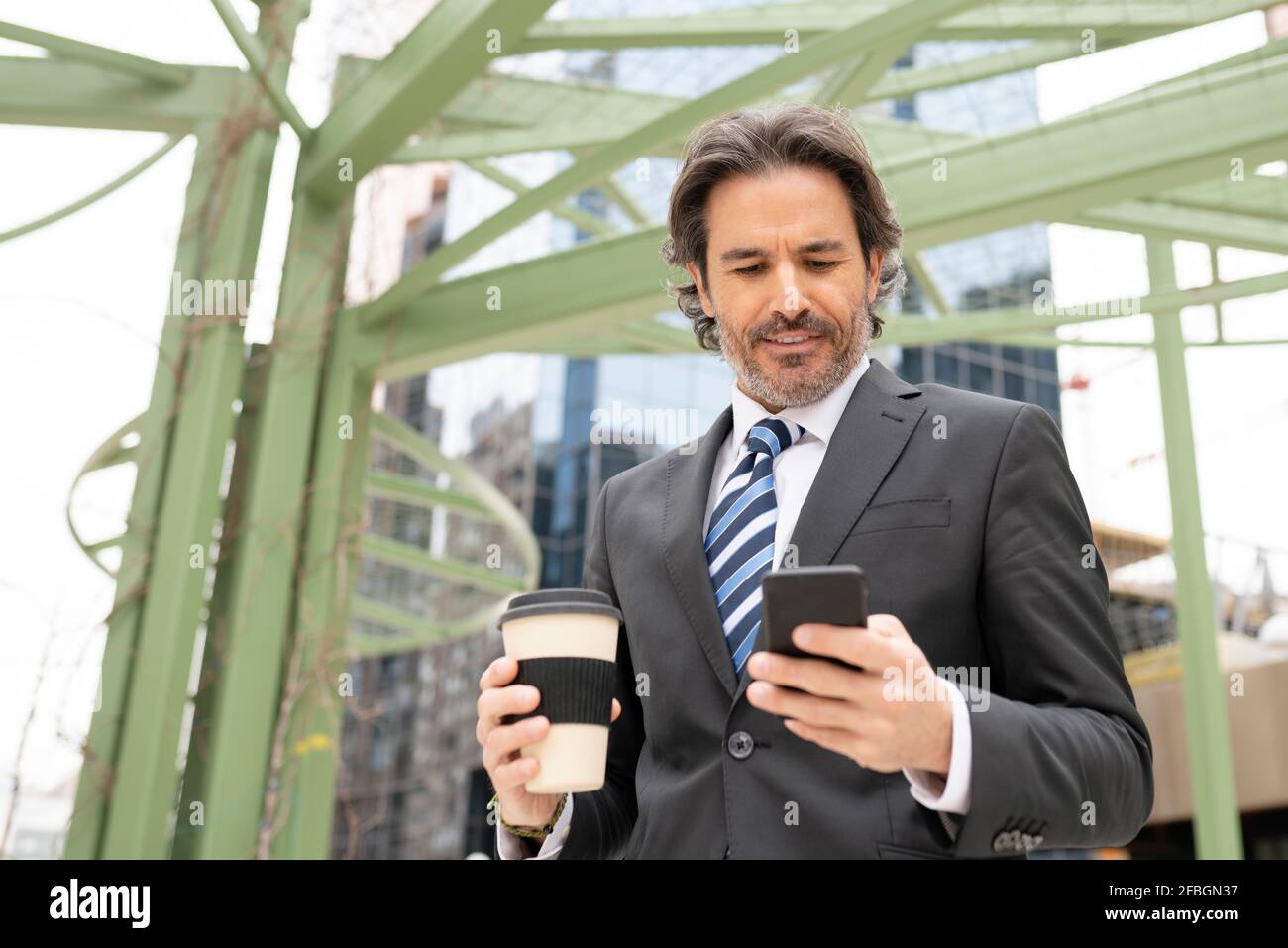 Uomo d'affari che tiene la tazza del caffè mentre usando il telefono mobile contro la struttura Foto Stock