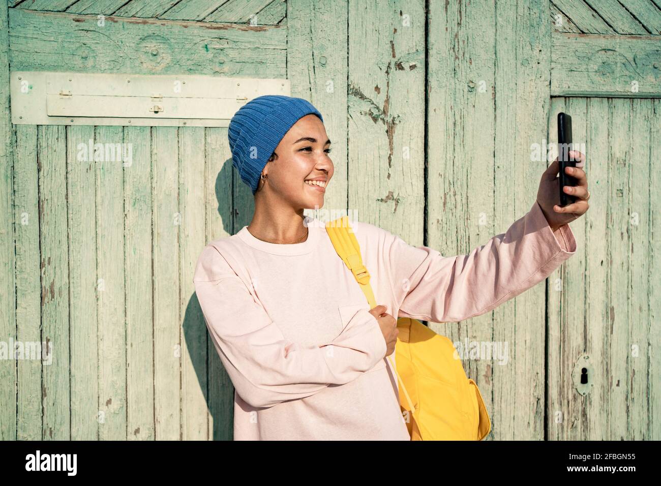 Donna felice in cappello a maglia che porta lo zaino mentre prende selfie tramite telefono cellulare davanti alla porta Foto Stock
