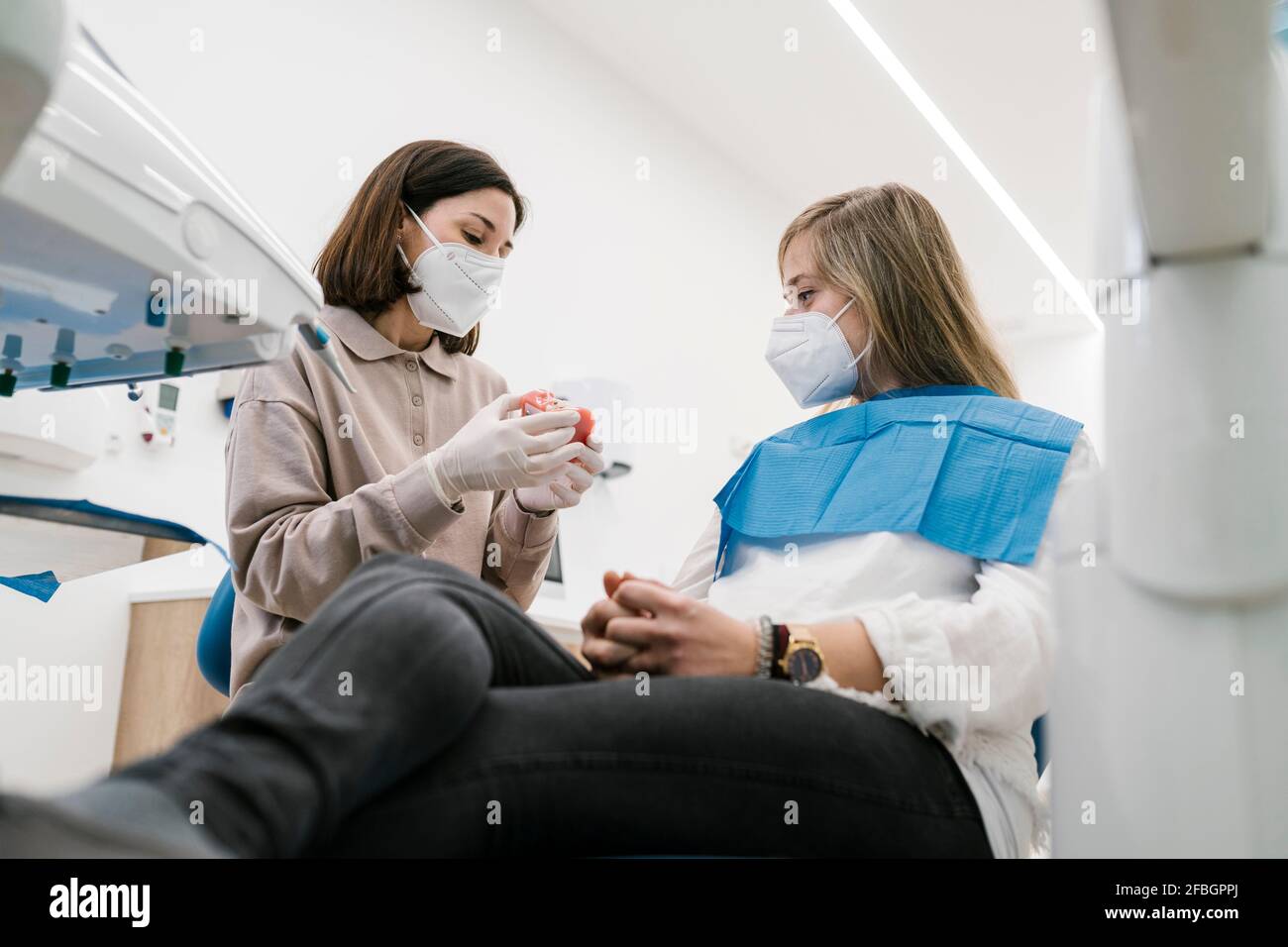 Ortodontista femminile indossando maschera facciale spiegare modello denti artificiali a. paziente in ufficio Foto Stock