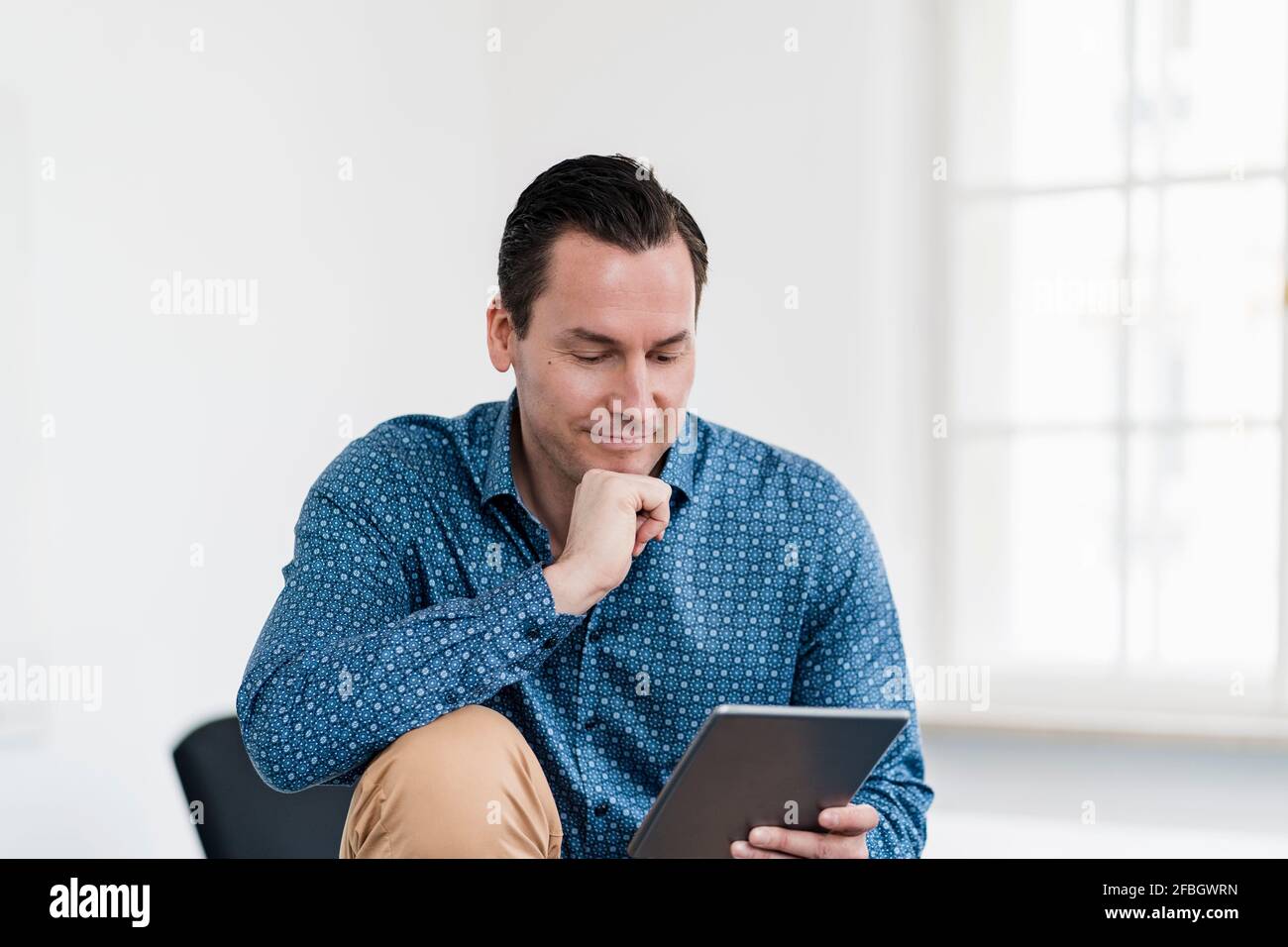 Uomo d'affari con la mano sul mento che usa il tablet digitale sul posto di lavoro Foto Stock