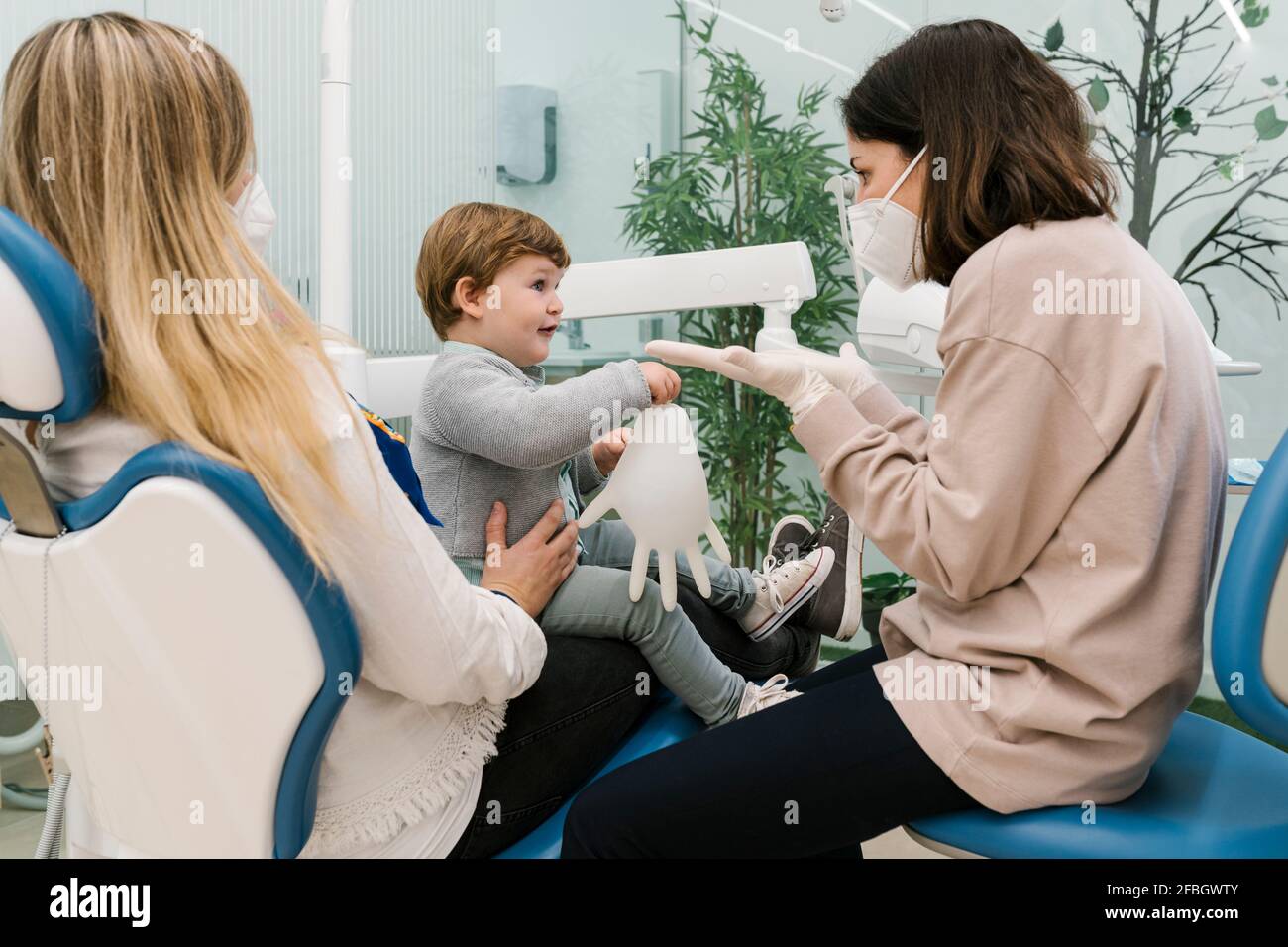 Dentista pediatrico che indossa una maschera protettiva che parla con il ragazzo seduto con madre in clinica Foto Stock