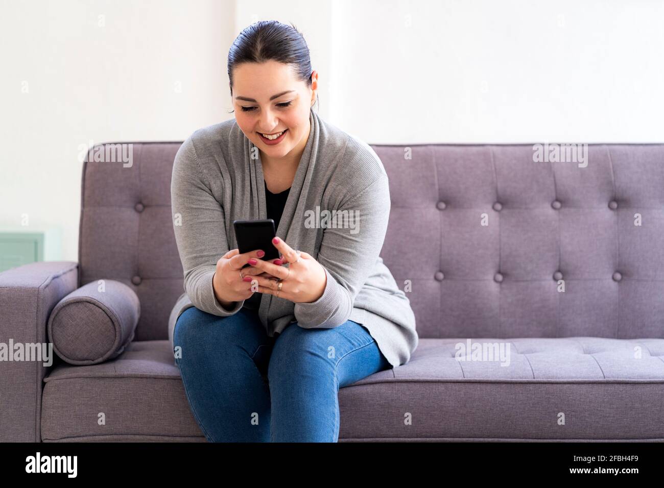 Donna sorridente che usa il telefono cellulare sul divano in soggiorno a casa Foto Stock