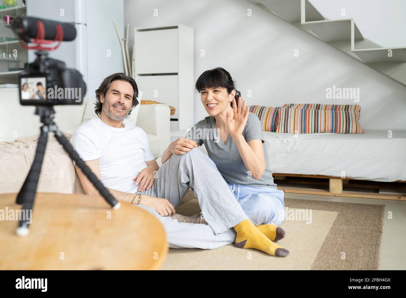 Donna sorridente che sventolava mentre si siede da un uomo che guarda la macchina fotografica a casa Foto Stock