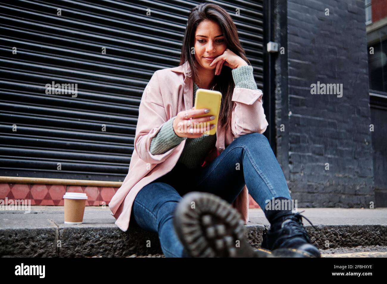 Bella donna con tazza da caffè utilizzando il telefono cellulare mentre si è seduti sul marciapiede Foto Stock