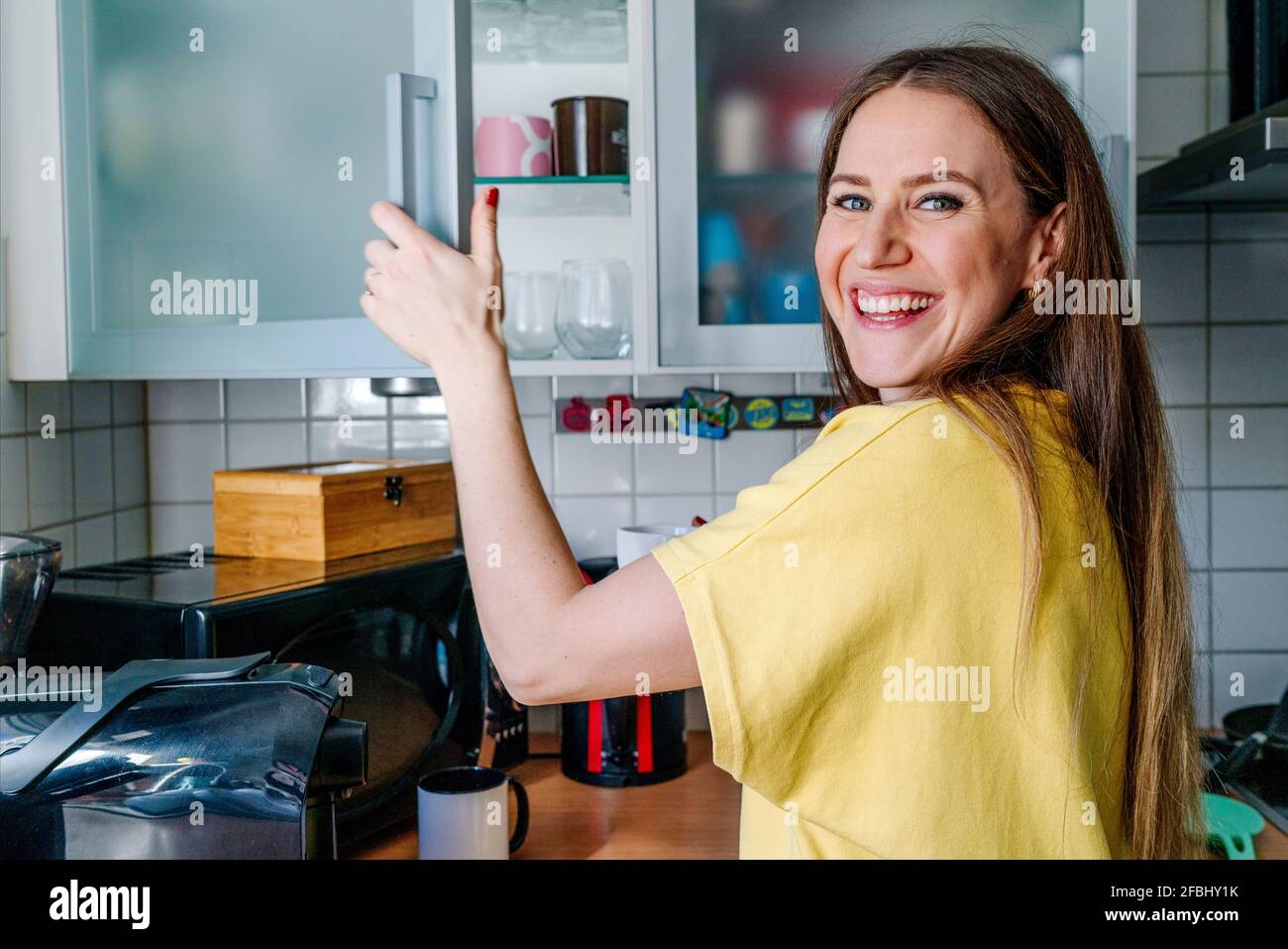 Donna felice con gli occhi grigi che aprono la porta del mobile dentro cucina a casa Foto Stock