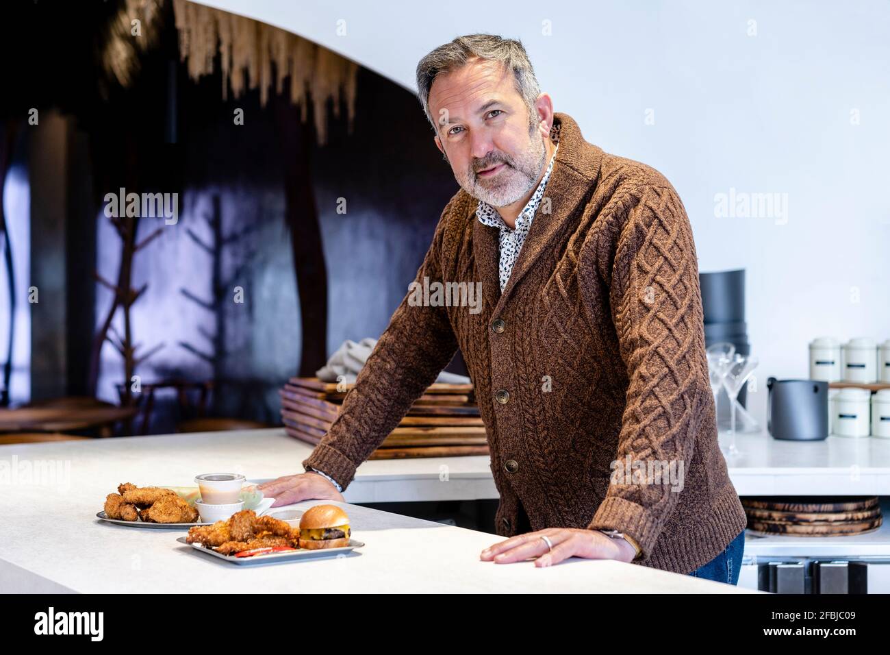 Uomo maturo in piedi al tavolo da cucina con piatto di hamburger e ali di pollo a casa Foto Stock