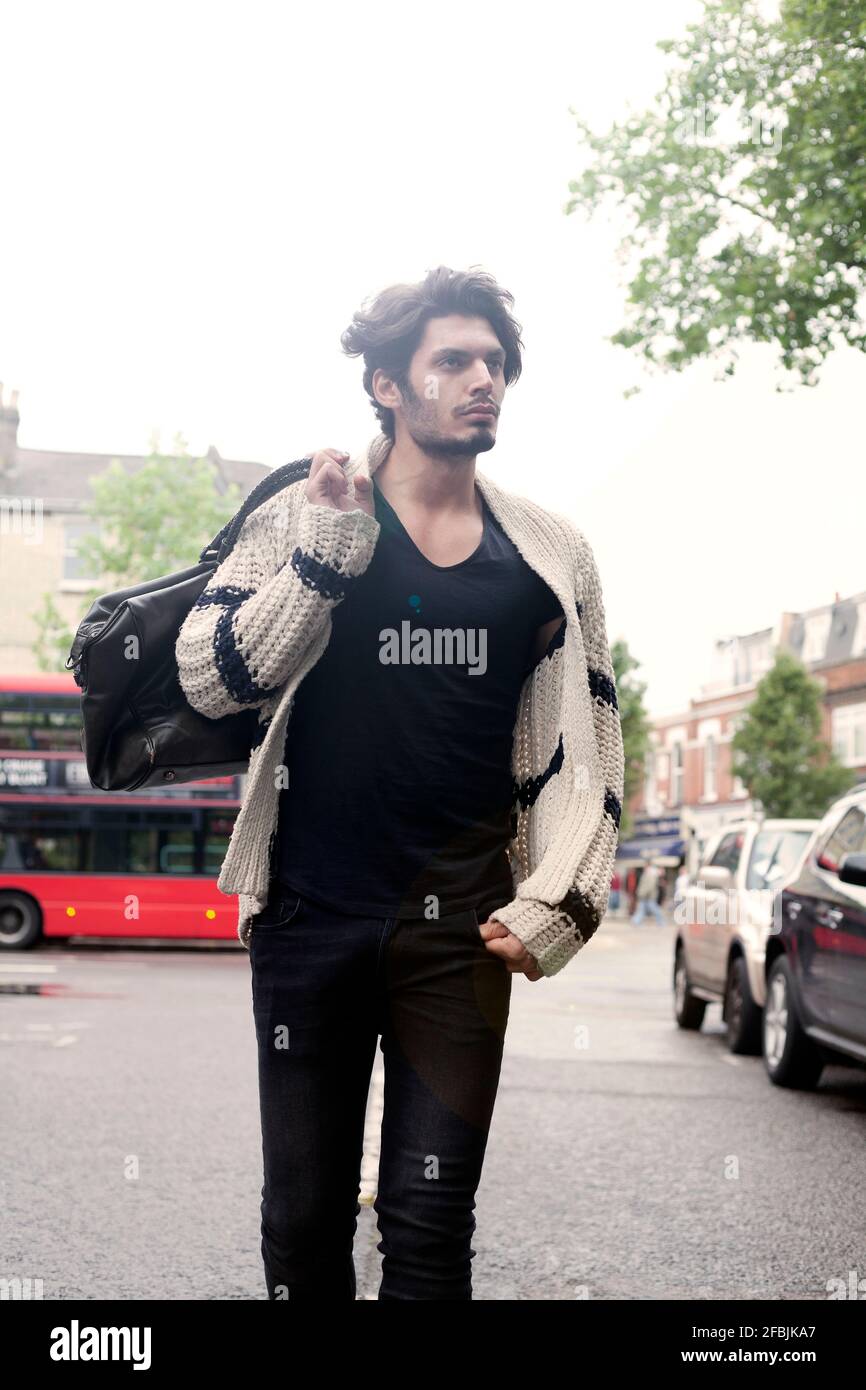 Ritratto di giovane bell'uomo che indossa cardigan camminando per strada con borsa in mano Foto Stock