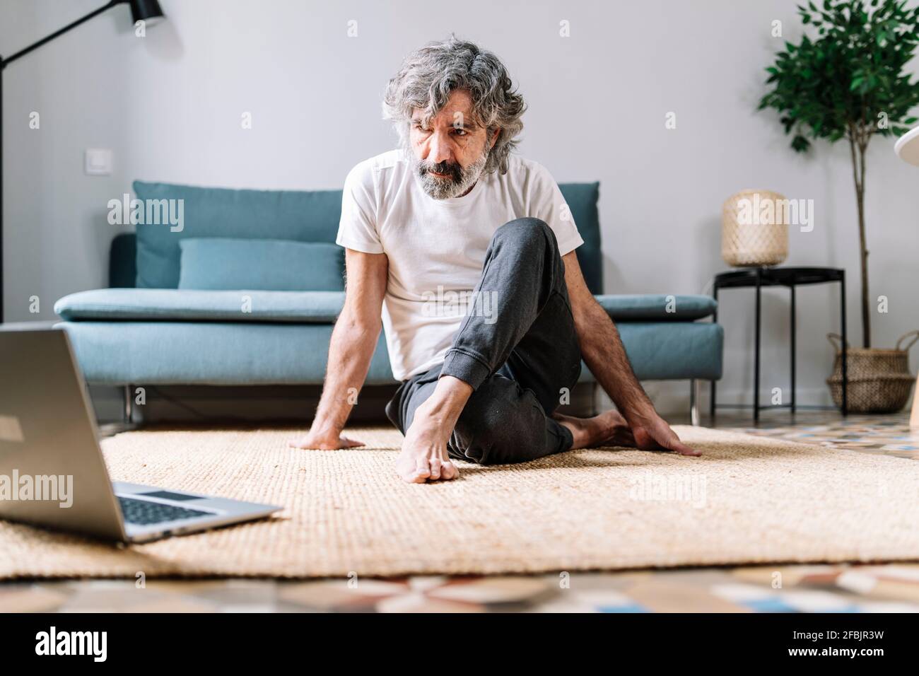 Uomo che fa yoga sulla moquette mentre guarda tutorial sul computer portatile a casa Foto Stock