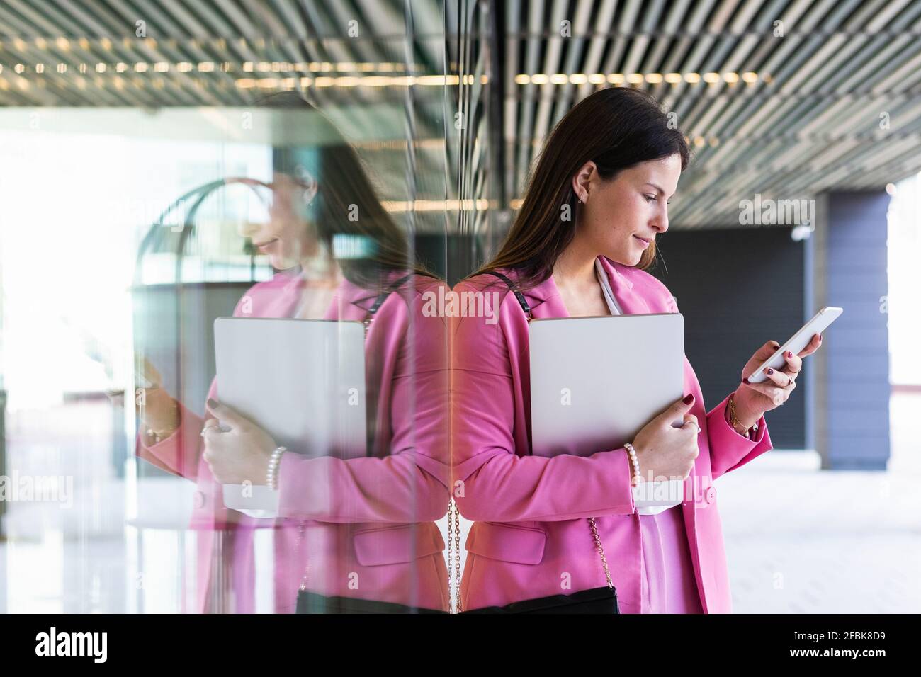 Giovane imprenditore femminile con computer portatile utilizzando il telefono cellulare mentre si appoggia su parete di vetro Foto Stock