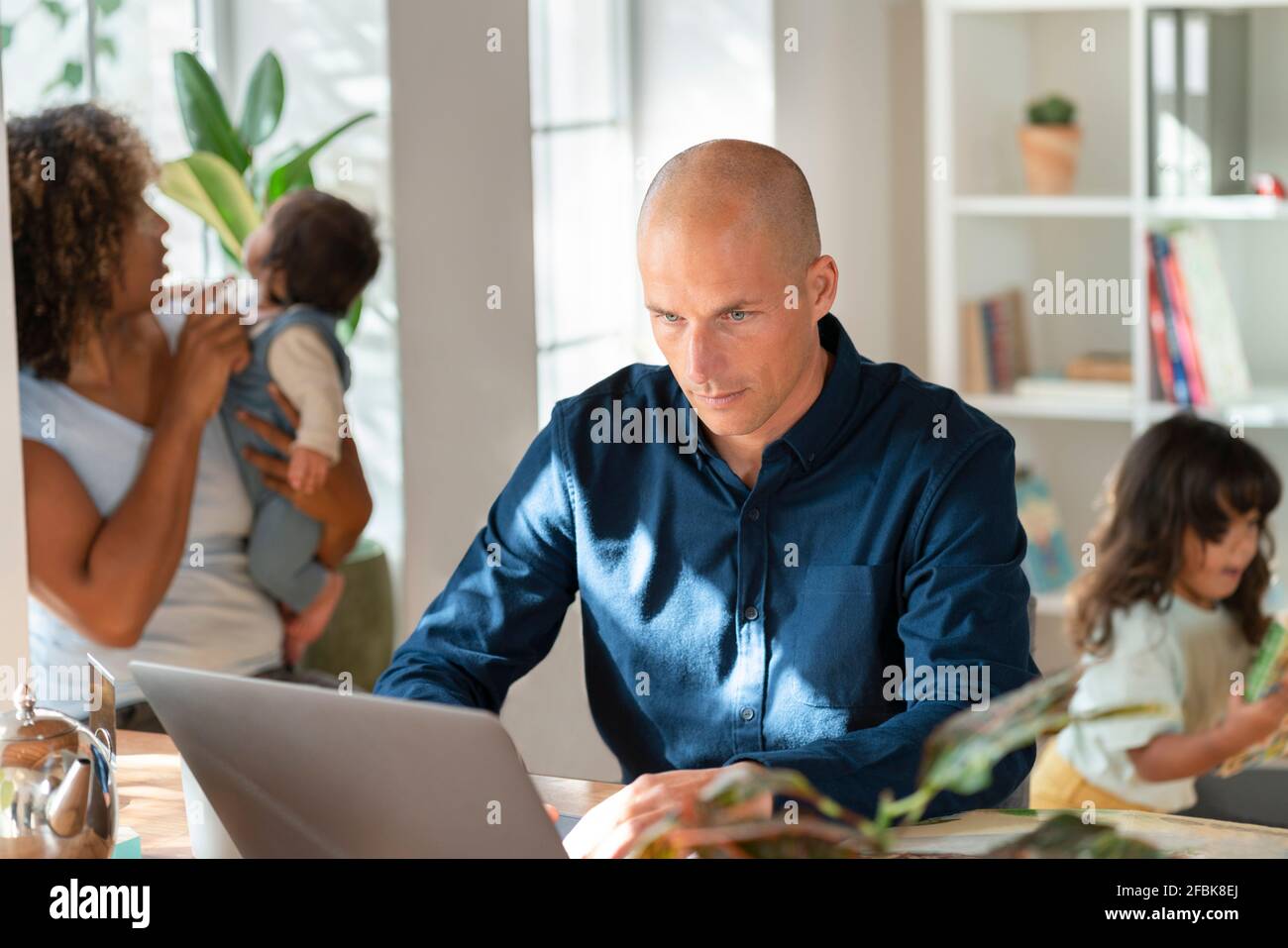 Padre che lavora su un computer portatile mentre la famiglia corre in background a. ufficio domestico Foto Stock