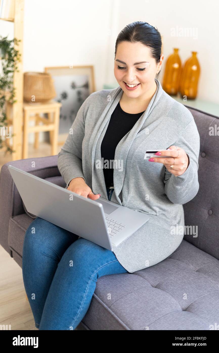 Donna sorridente che tiene la carta di credito mentre lo shopping in linea usando il laptop a casa Foto Stock