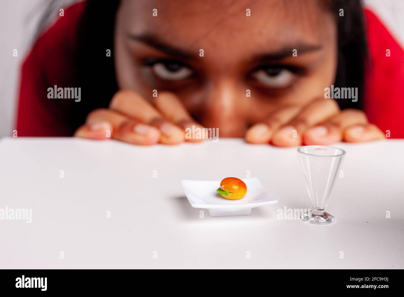 Giovane donna affamata che si intravea un invitante hamburger giocattolo in miniatura un piatto con i suoi occhi visibili sopra il tavolo in un divertente concetto di fast food Foto Stock