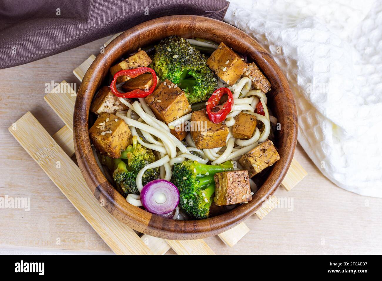 Vista dall'alto del tofu fritto vegetariano fatto in casa con tagliatelle e verdure (broccoli, pepe e cipolle) nella ciotola di legno sullo sfondo della cucina leggera Foto Stock