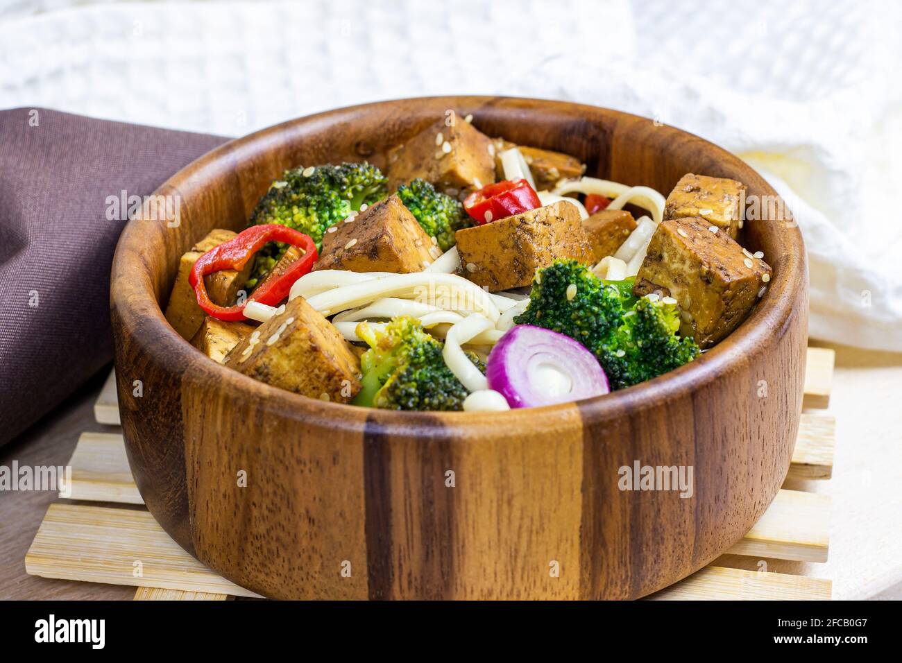 Tofu fritto vegetariano fatto in casa con tagliatelle e verdure (broccoli, pepe e cipolle) nella ciotola di legno su fondo cucina chiaro. Foto Stock