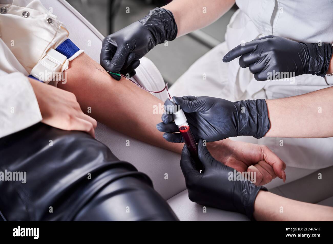 Chiudere la mano del medico con guanti neri che tengono la provetta con il sangue mentre l'assistente inserisce l'ago nel braccio del paziente. Scienziati di laboratorio medico che raccolgono campioni di sangue da giovane donna. Foto Stock