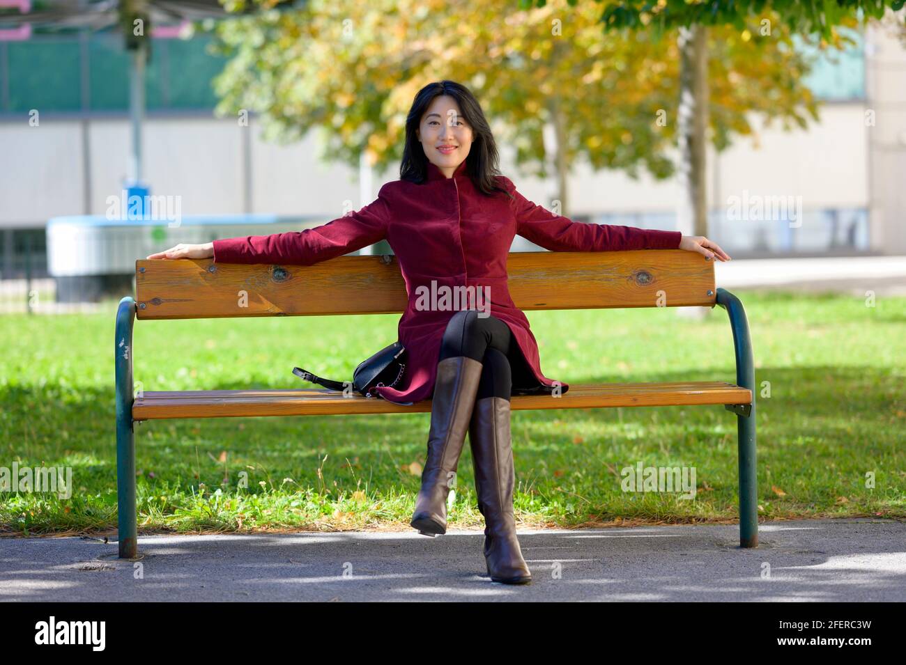 Bella donna cinese con lunghi capelli neri in una giornata di sole autunno in città, seduta su una panchina nel caldo sole d'autunno, Foto Stock