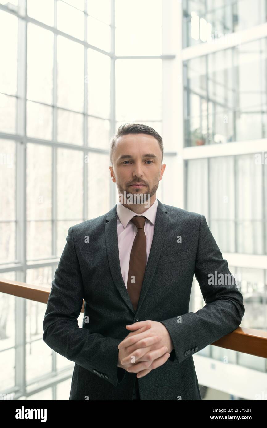 Ritratto di uomo d'affari serio bello con stoppie che indossa il vestito formale e cravatta appoggiata sulla ringhiera nel corridoio della sala d'affari Foto Stock